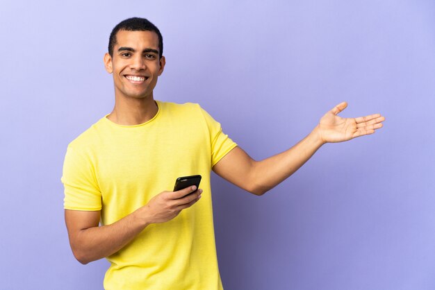 Homem afro-americano sobre parede roxa isolada usando telefone celular, estendendo as mãos para o lado para convidar para vir