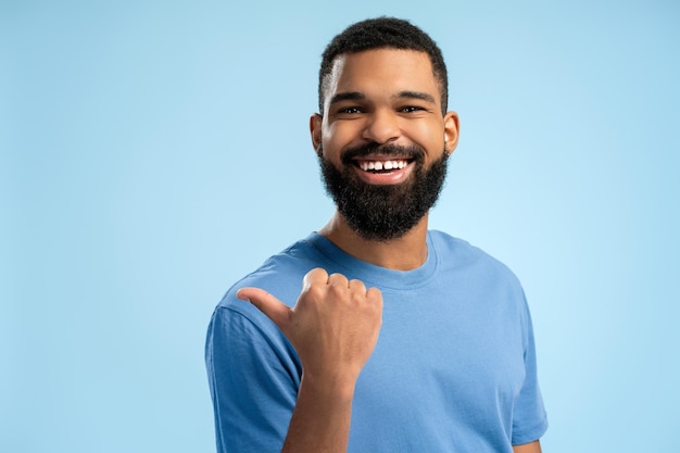 Homem afro-americano sorridente apontando para o lado com o polegar para cima sobre fundo azul com espaço de cópia