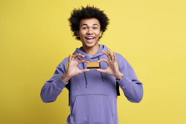 Homem afro-americano sorridente segurando o cartão de crédito isolado em fundo amarelo. compras, vendas