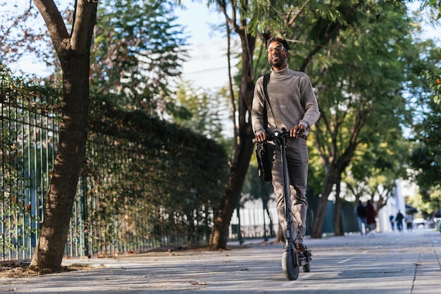 Homem afro-americano sorrindo com colete de pescoço alto movendo-se com sua scooter elétrica pela cidade