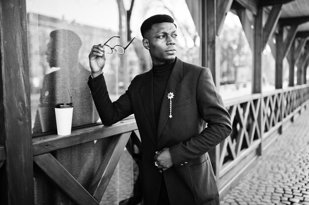 Homem afro-americano surpreendentemente vestindo blazer azul com broche de gola alta preta e óculos posou na rua cara negro elegante com xícara de café