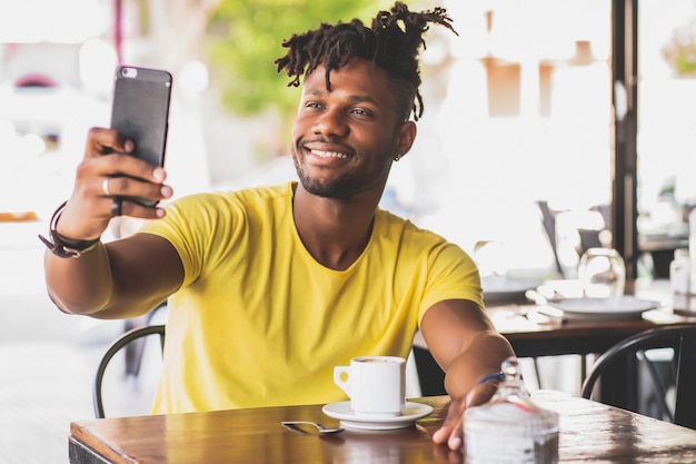 Homem afro-americano, tirando uma selfie com um telefone celular enquanto está sentado em uma cafeteria.