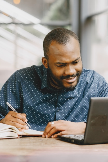 Homem afro-americano, trabalhando atrás de um laptop e escrevendo em um caderno. Homem com barba sentado em um café.