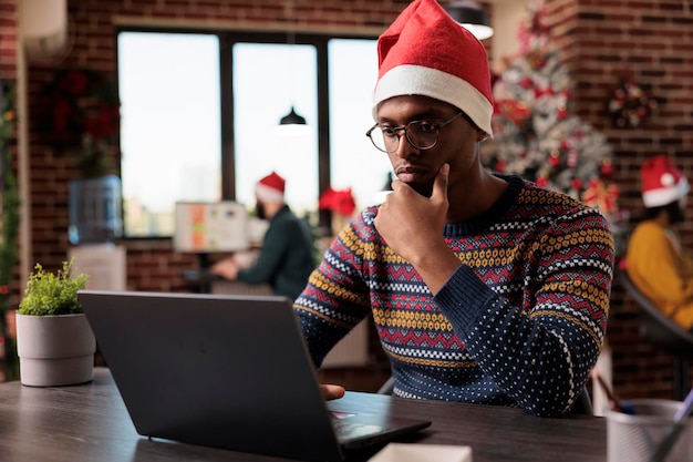 Homem afro-americano trabalhando no laptop no escritório, usando o computador no local de trabalho festivo decorado com árvore de natal e enfeites de férias. Trabalhador masculino no trabalho da empresa durante a temporada de inverno.