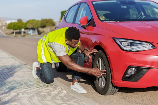 Homem afro-americano verifica o pneu de um carro vermelho com um colete amarelo