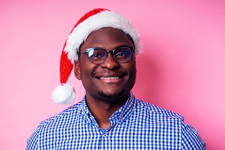 Foto homem afro-americano, vestindo elegante camisa xadrez grande sorriso no chapéu de papai noel com caixa de presente no fundo rosa studio. papai noel de pele escura parabenizando o feliz natal.
