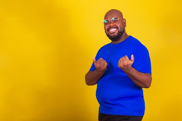 Homem afro-brasileiro bonito vestindo camisa azul de óculos em fundo amareloxAssinar venha venha venha aqui bem-vindo recepção receptiva recebendo gesto venha