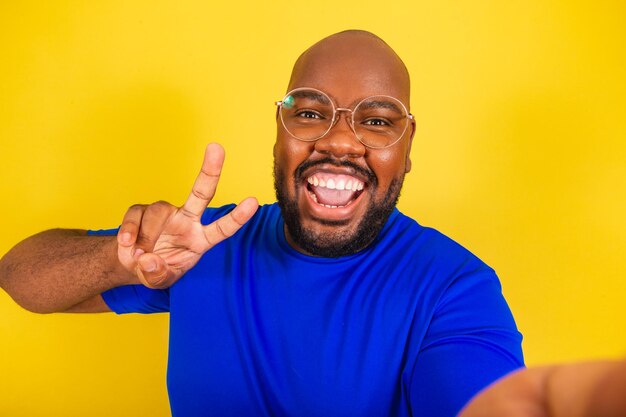 Homem afro brasileiro bonito vestindo camisa azul de óculos sobre fundo amarelo tomando selfie com sinal de paz e amor celular comunicação de chamada de vídeo foto interativa