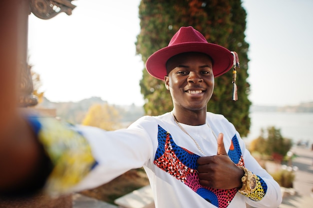 Homem afro-francês elegante e moderno de chapéu vermelho e roupa branca posou no dia do outono O modelo africano negro segura a câmera e faz selfie