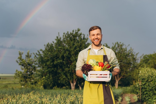 Homem agricultor feliz segurando cesta com legumes frescos no conceito de jardinagem de fundo de arco-íris e jardim