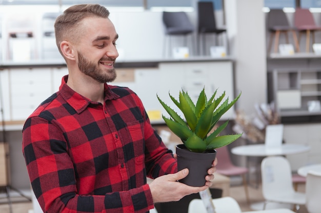 Homem alegre bonito sorrindo, segurando aloe em uma panela, comprando a decoração para casa na loja de móveis