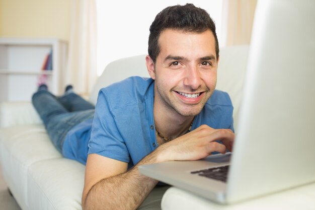 Homem alegre casual sentado no sofá usando o laptop olhando a câmera