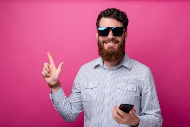 Homem alegre com barba no casual segurando smartphone e apontando para fora na copyspace