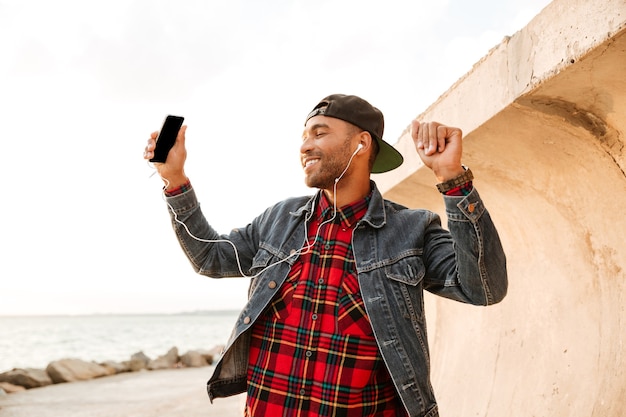 Homem alegre com boné, caminhando na praia enquanto ouve música com fones de ouvido.