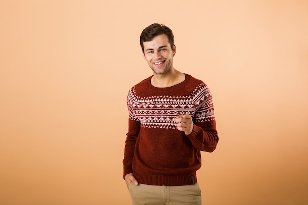 homem alegre com cerdas vestindo um suéter de tricô apontando o dedo para você, isolado sobre uma parede bege