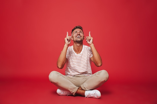 homem alegre de 30 anos em uma camiseta listrada apontando os dedos para cima, enquanto está sentado no chão com as pernas cruzadas isoladas no vermelho
