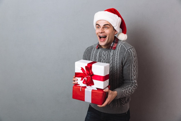 Homem alegre de suéter e chapéu de Natal segurando presentes enquanto desvia o olhar