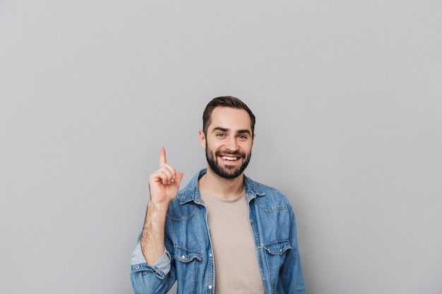 Homem alegre e animado vestindo uma camisa isolada na parede cinza, apontando para o espaço da cópia