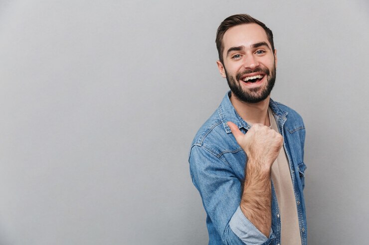 Foto homem alegre e animado vestindo uma camisa isolada na parede cinza, apontando para o espaço da cópia
