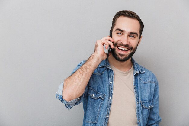Homem alegre e animado vestindo uma camisa isolada sobre uma parede cinza, falando no celular