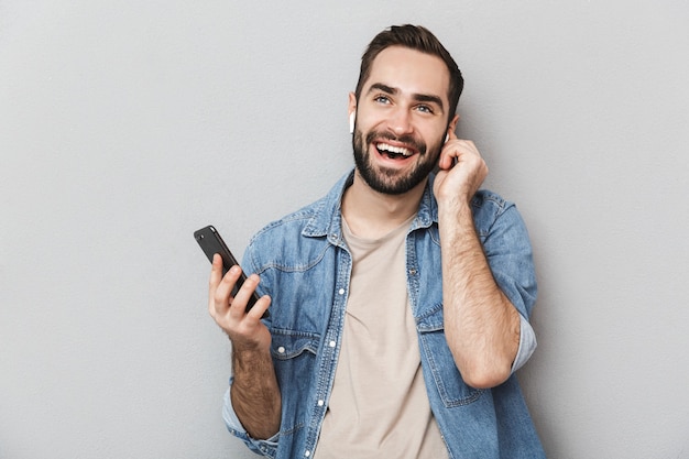 Homem alegre e animado vestindo uma camisa isolada sobre uma parede cinza, usando fones de ouvido, usando telefone celular