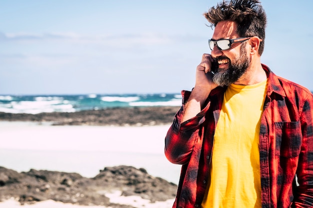 Homem alegre e feliz hippie com barba e óculos escuros falando ao telefone com amigos em atividades de lazer ao ar livre em beuaitufl lugar