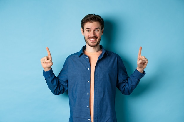 Homem alegre e sorridente em roupas casuais, apontando os dedos para cima e olhando para a câmera, mostrando o anúncio, de pé sobre fundo azul.