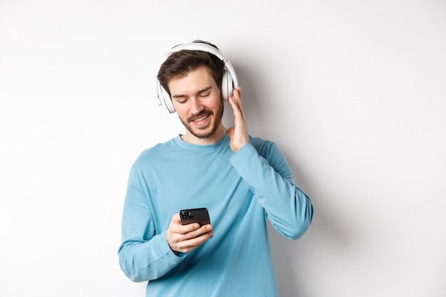 Homem alegre e sorridente, ouvindo música e olhando para o smartphone, lendo a mensagem no telefone, de pé no fundo branco.