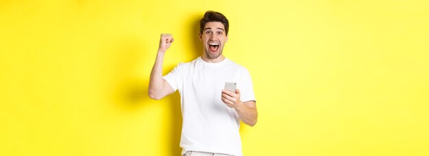 Foto homem alegre ganhando no smartphone levantando a mão e segurando o celular alcançando o objetivo do aplicativo em pé