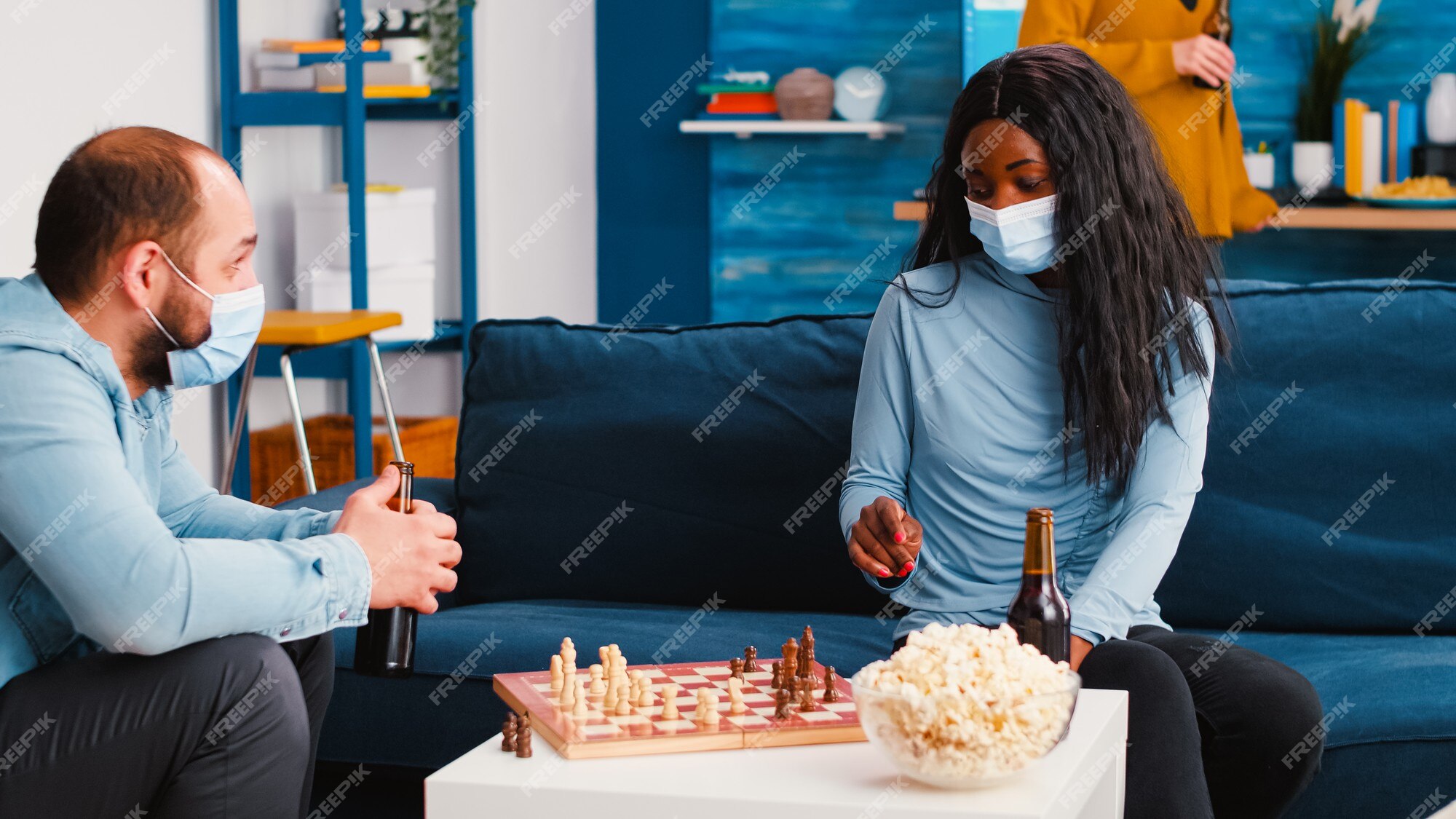 Homem alegre jogando xadrez com um amigo africano usando uma máscara contra  o covid 19, durante a pandemia global, sentado no sofá, bebendo cerveja e  comendo pipoca