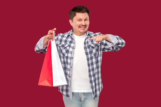 Homem alegre moderno de meia idade em camiseta branca e camisa quadriculada em pé e apontando o dedo para sacolas de compras e sorriso, olhando para a câmera. estúdio interno baleado em fundo vermelho isolado.