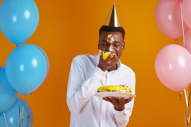 Homem alegre na tampa com rosto manchado, degustação de bolo de aniversário, fundo amarelo. homem sorridente tem surpresa, comemoração do evento, decoração de balões