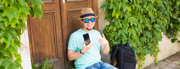 Homem alegre segurando e apontando o smartphone ao ar livre.