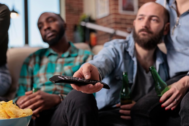 Homem alegre, trocando programas de canais de televisão com controle remoto de tv, procurando um bom filme ou filme de cinema. Pessoas se reunindo na festa em casa com garrafas de cerveja e pizza. Fechar-se.