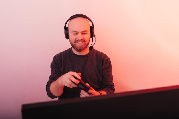 Homem alegre usando um fone de ouvido está brincando com seu joystick no computador Foto feita com luz vermelha