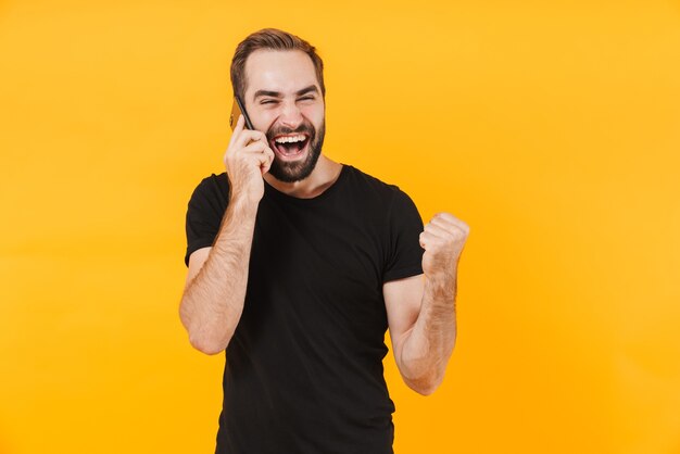 Homem alegre vestindo camiseta preta básica, regozijando-se e falando no smartphone isolado sobre a parede amarela