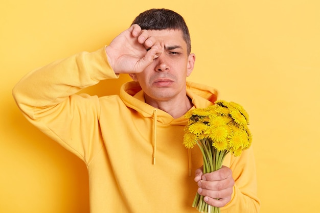 Homem alérgico doente doente vestindo capuz estilo casual esfregando os olhos sofre de sintomas de alergia ao pólen febre alta segurando flor florescendo posando isolado sobre fundo amarelo