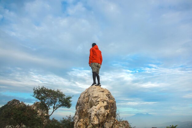 homem alpinista com jaqueta laranja em cima de pedra e desfrutando de uma vista incrível no céu azul brilhante