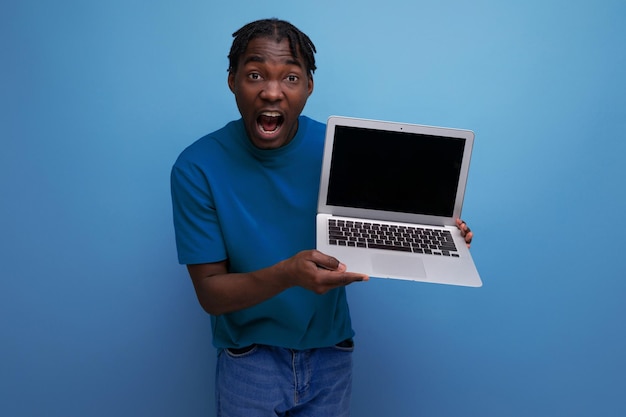 Homem americano positivo mostrando a tela do laptop no fundo do estúdio com maquete e espaço para cópia