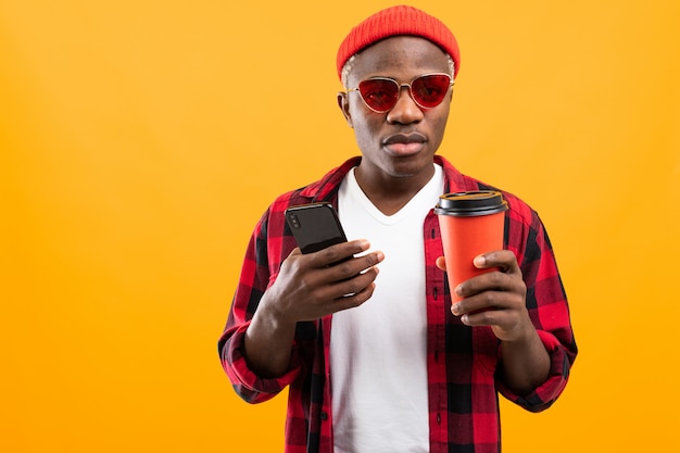 Homem americano preto bonito com telefone e copo de café para viagem em fundo amarelo studio