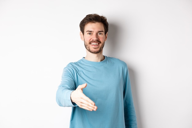 Homem amigável e sorridente estende a mão, diz olá e dá o braço para um aperto de mão, em pé sobre um fundo branco