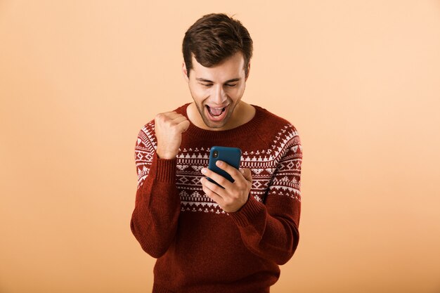 homem animado com a barba por fazer usando um suéter de tricô usando smartphone, isolado sobre uma parede bege