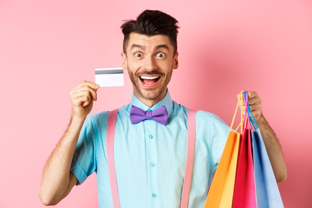 Homem animado mostrando cartão de crédito de plástico e sacolas de compras, sorrindo espantado ao comprar presentes, em pé na rosa.