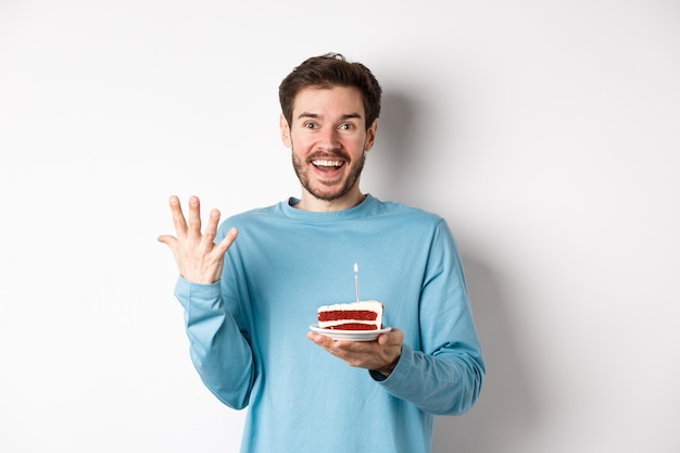 Homem animado recebe surpresa de aniversário, segurando um bolo de aniversário e sorrindo feliz, em pé sobre um fundo branco, fazendo um pedido em uma vela acesa.