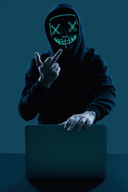 Foto homem anônimo em um capuz preto e máscara de néon invadindo um computador