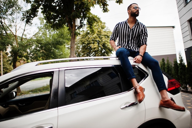 Homem árabe bem sucedido usa camisa listrada e óculos de sol sentados no telhado de seu carro suv branco.