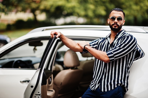 Homem árabe bem-sucedido usa camisa listrada e óculos escuros posar perto de seu carro suv branco. homens árabes elegantes no transporte.