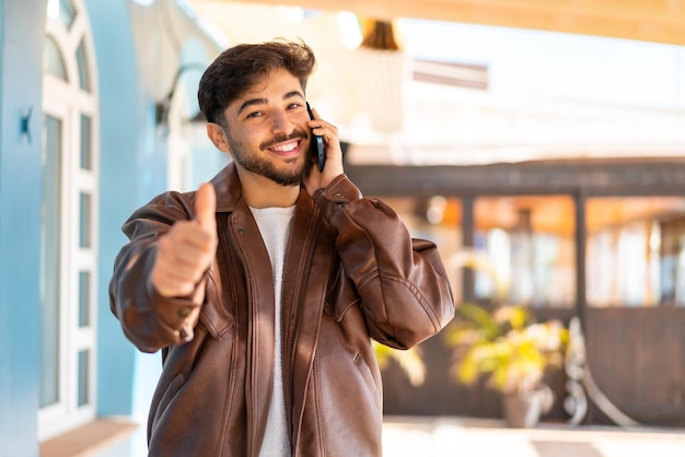 Homem árabe bonito ao ar livre, mantendo uma conversa com o celular enquanto faz sinal de positivo