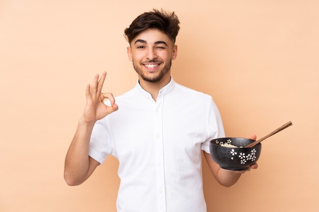 Homem árabe bonito isolado em uma parede bege mostrando um sinal de ok com os dedos enquanto segura uma tigela de macarrão com pauzinhos