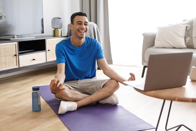 Homem árabe calmo positivo em roupas esportivas sentado em pose de lótus tendo aula de ioga online meditando perto de laptop em casa